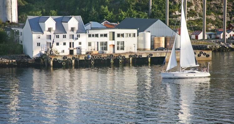 White sailboat glides past a waterfront warehouse complex reflecting in calm fjord water.