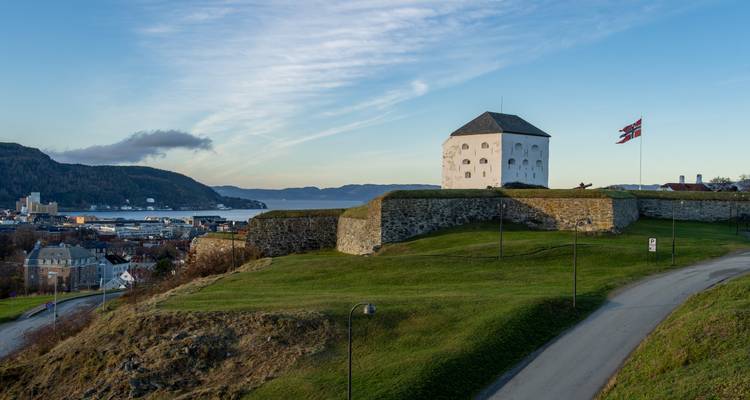 Kristiansten Fortress overlooks Trondheim and its fjord under a blue morning sky with Norwegian flag waving.