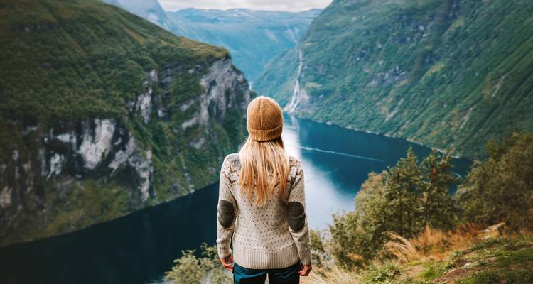 Traveler in wool sweater and beanie gazes over the dramatic blue Geirangerfjord from a high cliff edge.