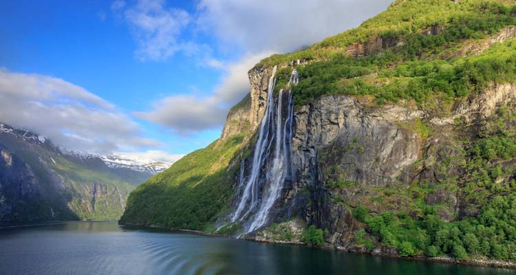Tall waterfall plunges over a green cliff into a calm fjord with blue skies and scattered clouds.