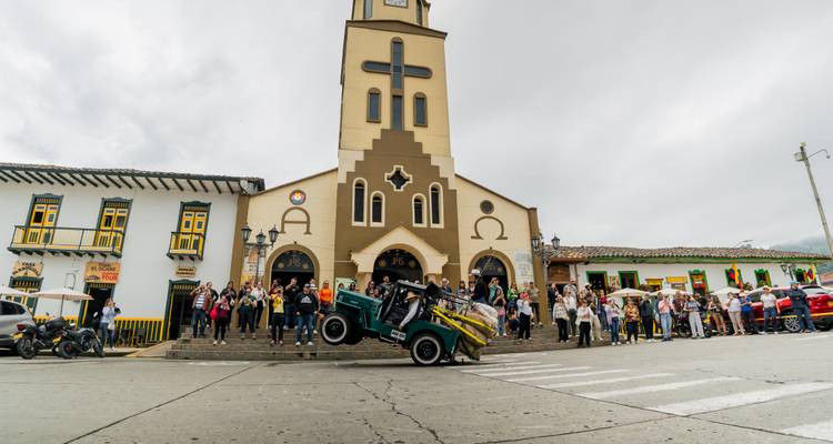 Un Jeep vert soulève ses roues avant devant une église beige tandis qu'une large foule observe depuis les marches de la place.