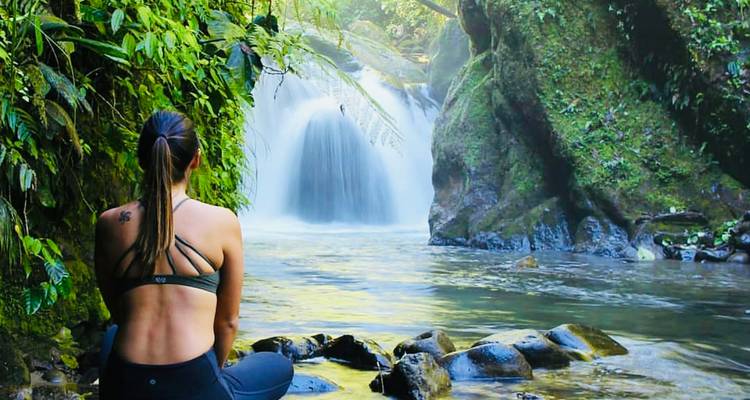 Young woman in sportswear sits on riverside rocks admiring a jungle waterfall cascading into a tranquil pool.