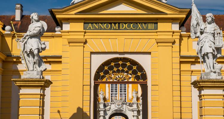 Puerta amarilla barroca de la Abadía de Melk adornada con estatuas e inscripción latina bajo un cielo azul.
