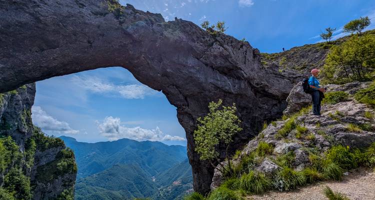 Une arche de pierre naturelle encadre un randonneur se tenant sur un rebord herbeux surplombant un vaste paysage montagneux.