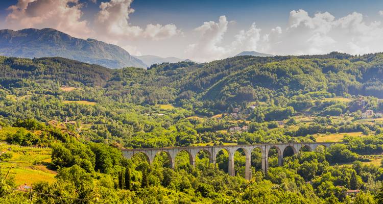 Un long viaduc ferroviaire en pierre enjambe une vallée verdoyante entourée de collines boisées ondulantes sous un ciel partiellement nuageux.