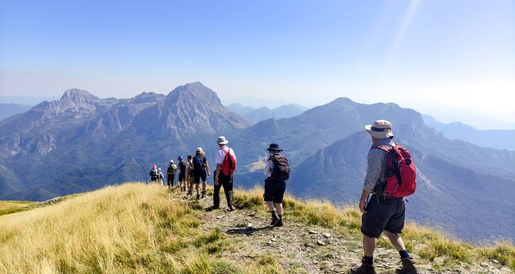 Une file de randonneurs avec des sacs à dos suit une crête herbeuse étroite en direction de sommets rocheux imposants sous un ciel bleu brumeux.