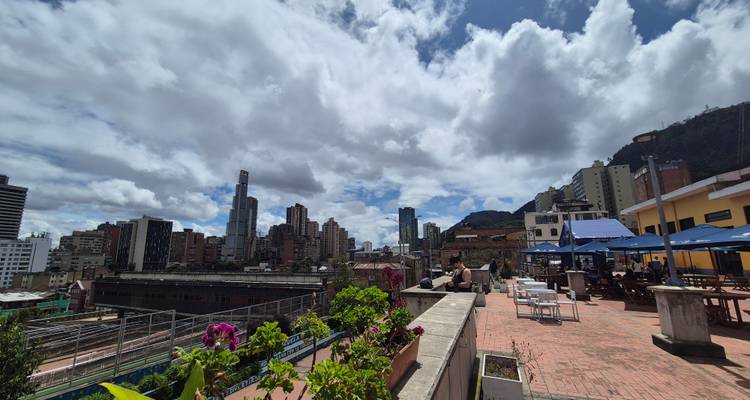 Vue panoramique depuis une terrasse d'une ville avec des gratte-ciel, des tables de patio et des fleurs en pot sous un ciel partiellement nuageux.