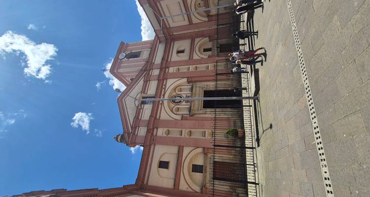 Façade d'église rose et crème imposante avec clocher à horloge sous un ciel bleu éclatant, quelques visiteurs se tenant sur la place en pierre devant.