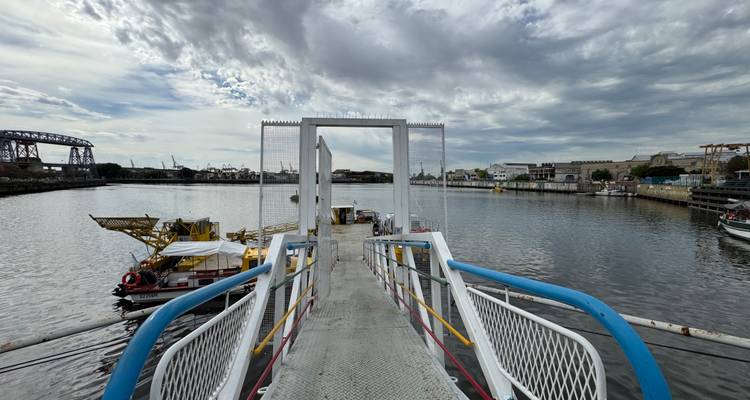 Metalen loopbrug naar een drijvende pontons op een brede stadskanaal onder een textuurrijke wolkendek.