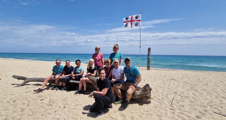 Smiling tour group poses on a driftwood log on a wide sandy beach with turquoise sea and the Sardinian flag fluttering behind.