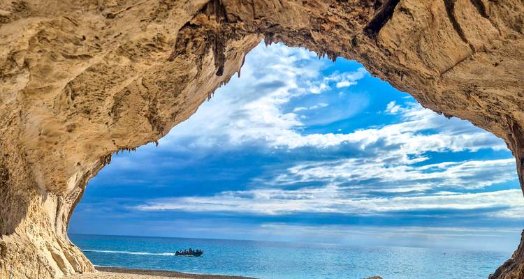 View from inside a seaside cave framing bright turquoise water, blue sky and a small motorboat in the distance.