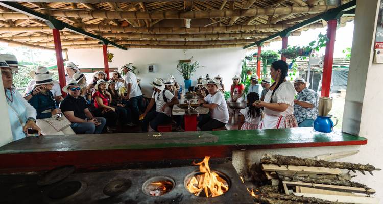 Visitors seated inside an open-air country kitchen while a host demonstrates cooking over a wood fire.
