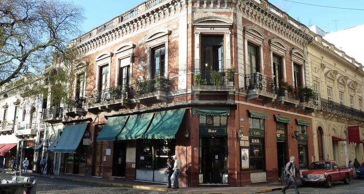 Bâtiment historique d'angle à deux étages avec des auvents verts et des balcons sur une rue pavée dans un quartier animé.
