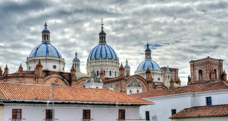 Blue and white domes of Cuenca Cathedral rise above red-tiled rooftops against a textured cloudy sky.