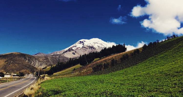 Paved Andean road leads toward a snow-capped Chimborazo peak framed by green hills and deep blue sky.