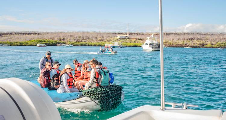 Tourists wearing life jackets ride an inflatable zodiac in turquoise Galapagos waters with yachts in background
