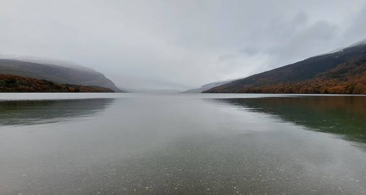 Vista simétrica hacia abajo de un lago tipo fiordo brumoso entre montañas oscuras bajo nubes bajas.