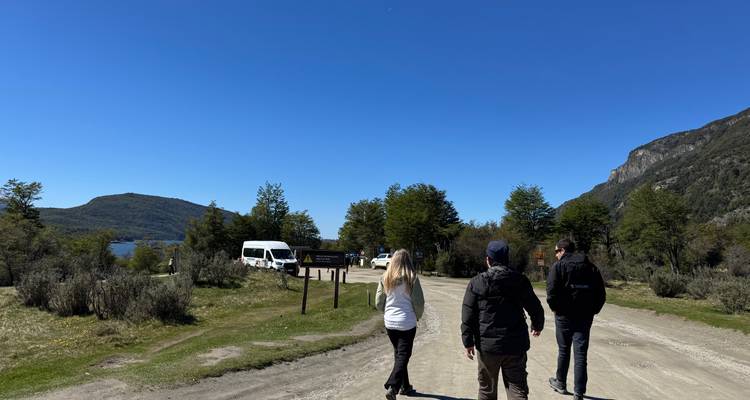 Tres viajeros caminan por un camino de tierra junto a un lago con furgonetas de tour y colinas boscosas alrededor.