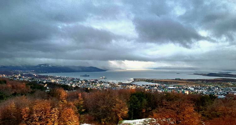 Vista panorámica sobre la ciudad de Ushuaia y el Canal Beagle enmarcados por nubes dramáticas y bosques de otoño.