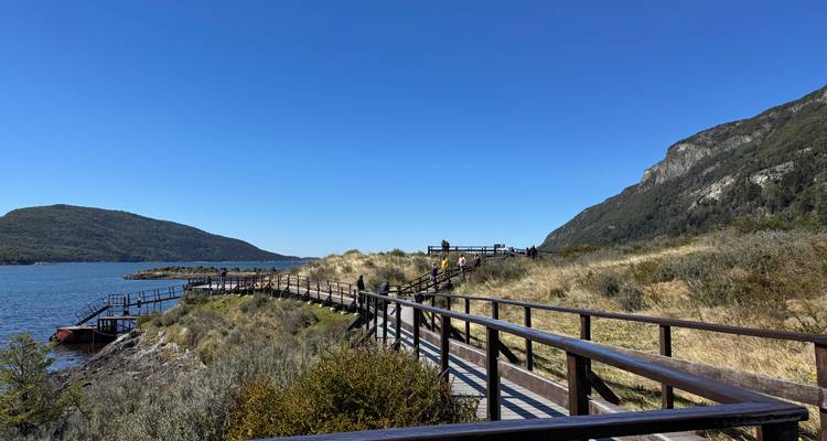 Un elevado paseo de madera serpentea a lo largo de la orilla del lago bajo colinas verdes y cielos azules, ofreciendo puntos de vista panorámicos.