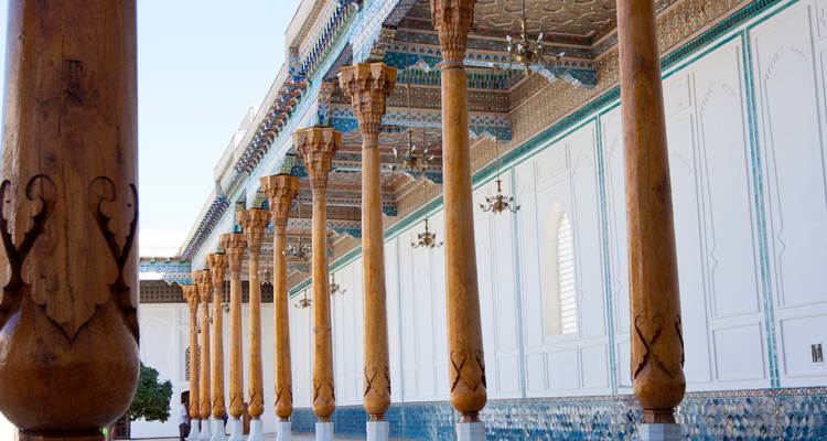Row of elaborately carved wooden columns supporting a richly decorated ceiling of a historic Uzbek palace.