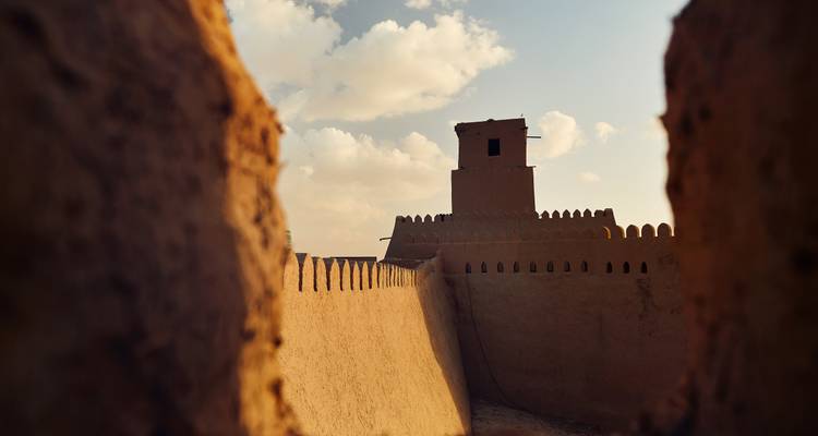 Adobe fort tower framed between two mud walls lit by warm late afternoon light.
