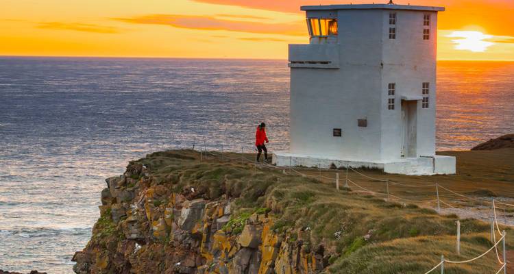 Un pequeño faro blanco encaramado en un acantilado cubierto de hierba al atardecer vivido mientras un visitante solitario admira la vista.