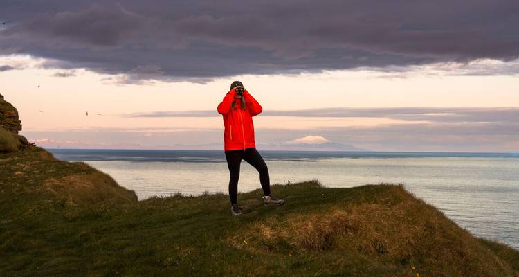 Un senderista con una chaqueta roja brillante está de pie en el borde de un acantilado cubierto de pasto tomando fotografías sobre el océano al atardecer.