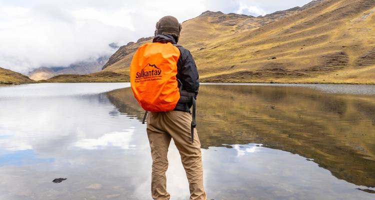 Persona parada junto a un lago reflectante, sosteniendo un bastón de senderismo.