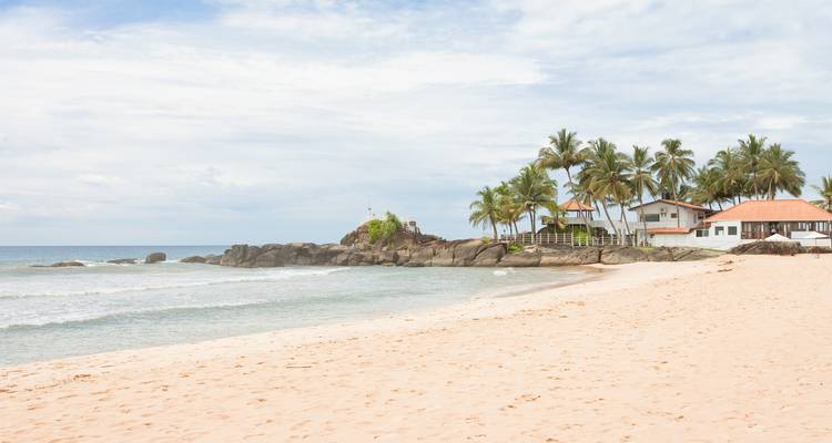 Ruhiger Strand mit Palmen und einem Haus auf den Felsen.