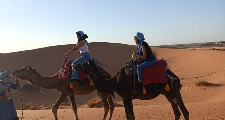 Dos mujeres montan camellos guiados por un guía vestido de azul sobre las ondulantes dunas del Sahara.