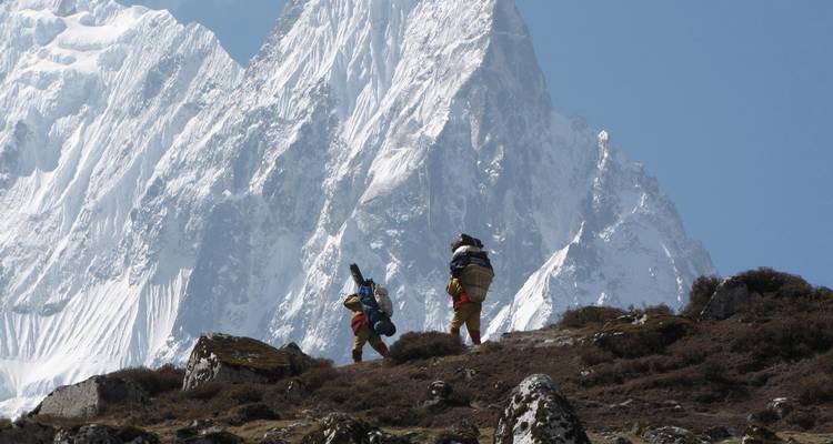 Porters carrying loads uphill with massive snowy peaks in the background.