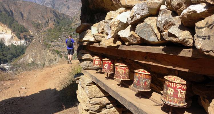 A hiker on a trail with prayer wheels and a mountain backdrop.