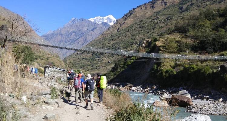 Randonneurs marchant le long d'une rivière avec un pont de corde et un décor de montagne.