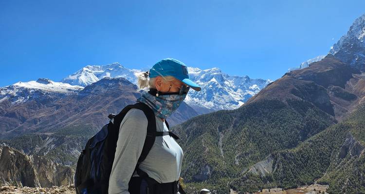 Un trekista enmascarado con mochila se detiene contra un telón de fondo espectacular de laderas boscosas y altos picos nevados.