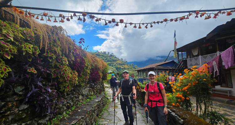Los excursionistas pasean por un callejón del pueblo bordeado de flores coloridas bajo nubes de monzón dramáticas y espacios azules.