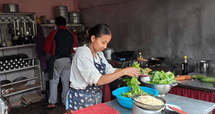 Un cocinero prepara verduras frescas en una modesta cocina de montaña mientras otra persona trabaja de fondo.