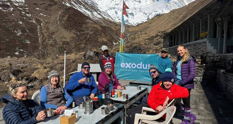 Wanderer entspannen sich mit heißen Getränken an einem Tisch im Freien eines Outdoor-Cafés unter schneebedeckten Bergen und einem Exodus-Banner.