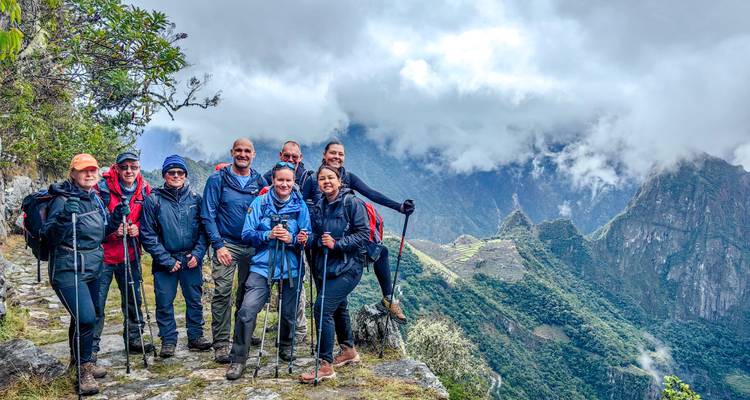 Ein fröhliches Trekking-Team versammelt sich an einer Felskante mit wolkenumhüllten Ruinen darunter.