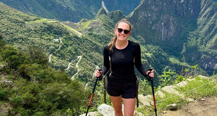 Solo trekker with poles smiles with Machu Picchu terraces visible far below.