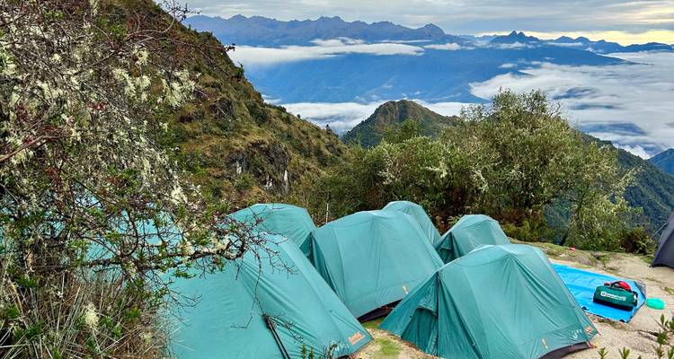 Row of teal expedition tents on a high mountain ledge above cloud-filled valleys.
