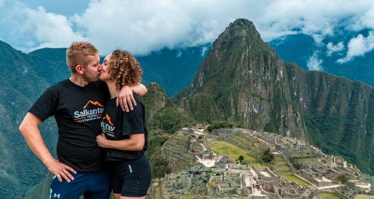 Ein Paar in Salkantay-Trek-Shirts küsst sich an einem Aussichtspunkt mit Blick auf Machu Picchus antike Terrassen und Gipfel.