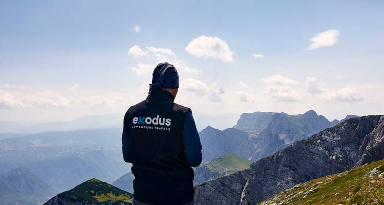 Ein Trekker in einer Mütze und einem Westen blickt über dramatische alpine Grate und Täler unter einem strahlend blauen Himmel.