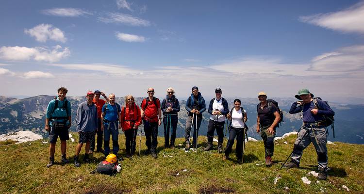 Ein vollständiges Trekking-Team posiert mit Stöcken auf einem offenen, grasbewachsenen Gipfel mit dunstig erscheinenden Gipfeln hinter ihnen.