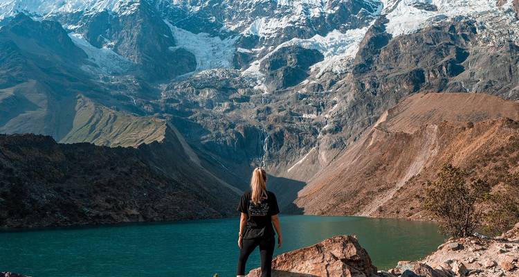 Solo traveller stands on rocks overlooking emerald Humantay Lake beneath towering glaciers.