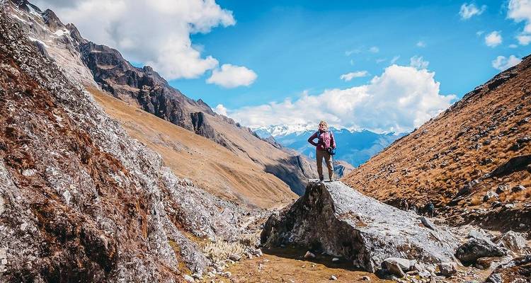 Hiker stands triumphantly atop a boulder in a high alpine valley under bright blue skies.