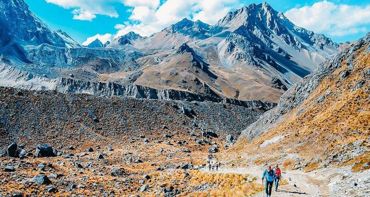Trekker group winds along a rugged mountain trail beneath jagged peaks and blue skies.