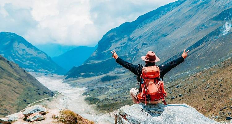 Backpacker with arms raised sits on cliff edge overlooking a dramatic Andean valley.