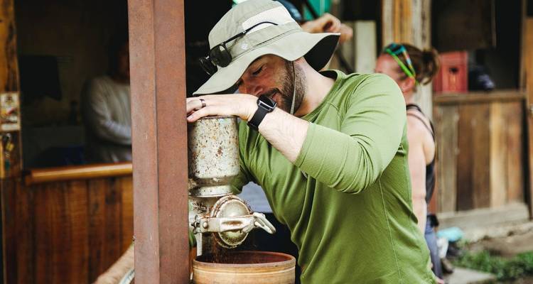 Traveller in sun hat grinds coffee beans using a rustic open-air grinder during a farm visit.