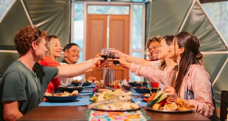 Happy trekkers seated around a table inside a geodesic dome clinking glasses over a colourful meal.